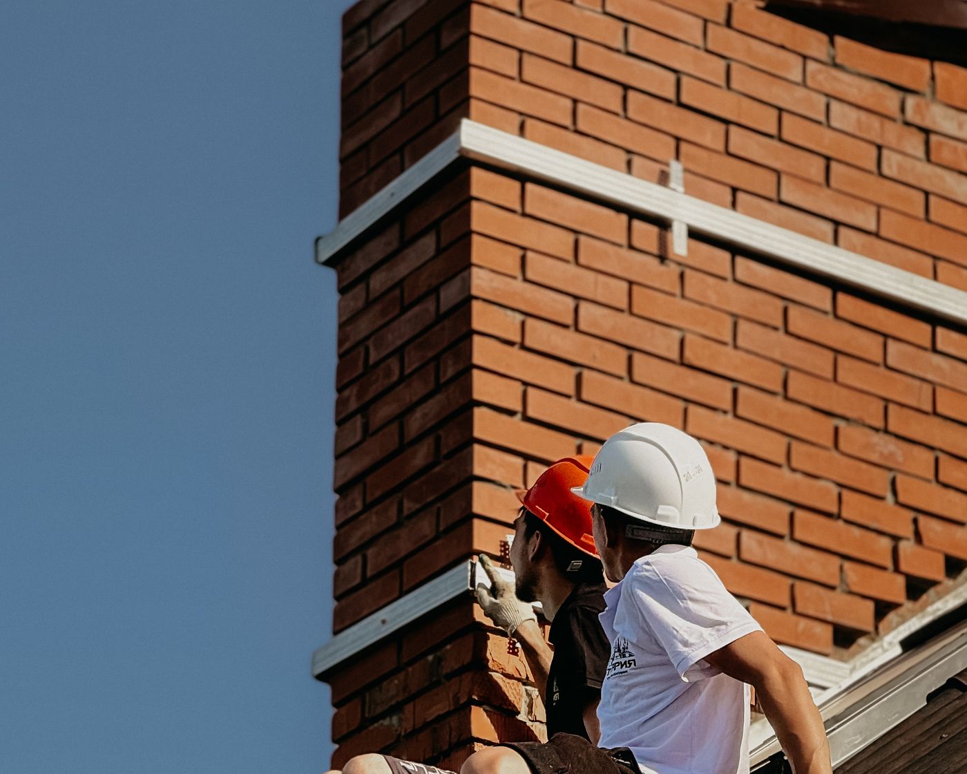 Two workers in hard hats at a brick chimney on a rooftop