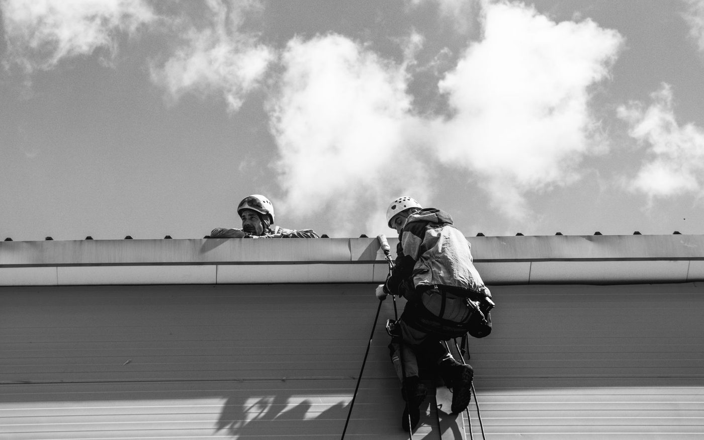Two workers in safety gear at the edge of a flat roof, in black and white