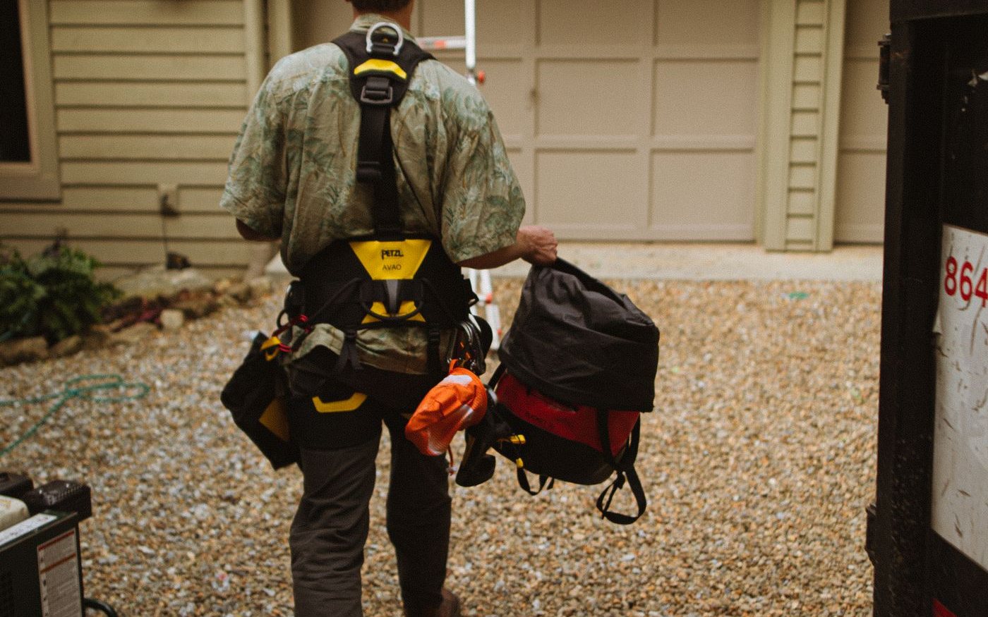 Roofer in a harness carrying tools toward a ladder at a house
