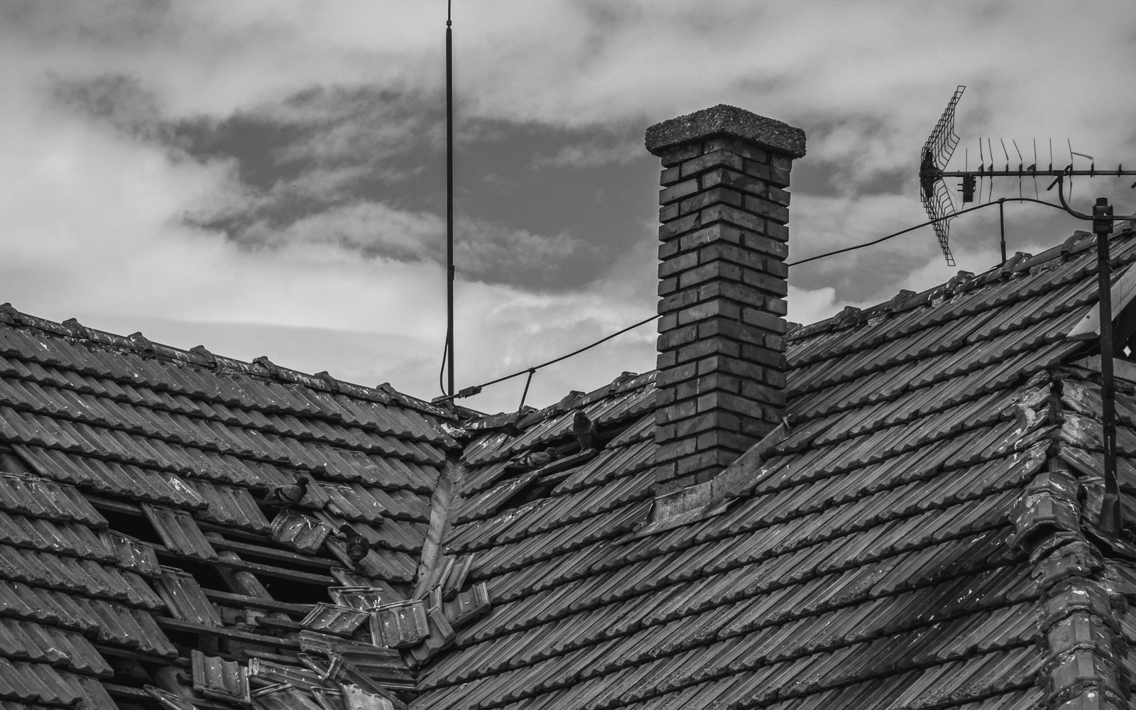 Storm-damaged tile roof broken open beside a chimney, in black and white